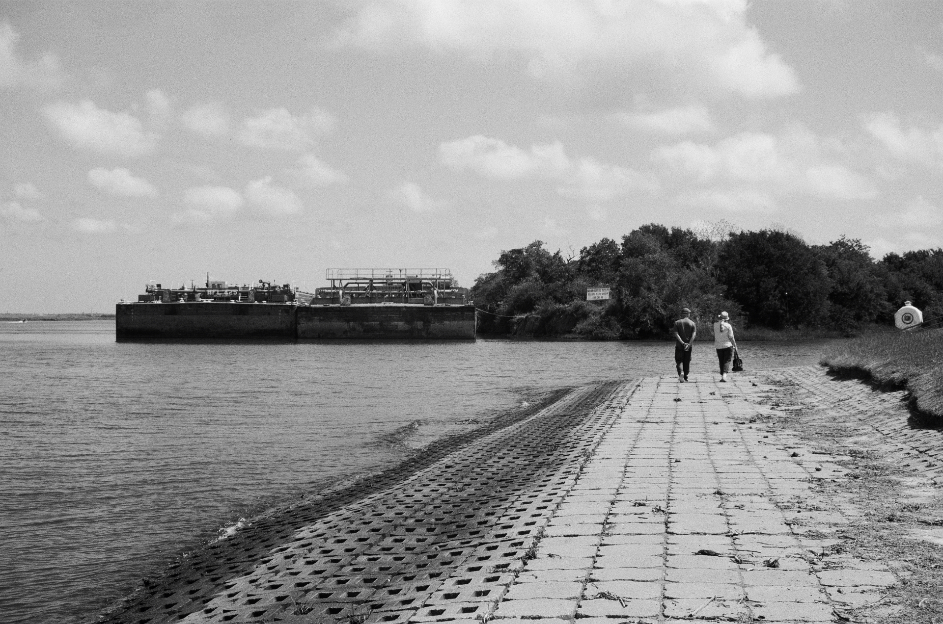 Two people walking along a sloped riverfront beside an industrial barge